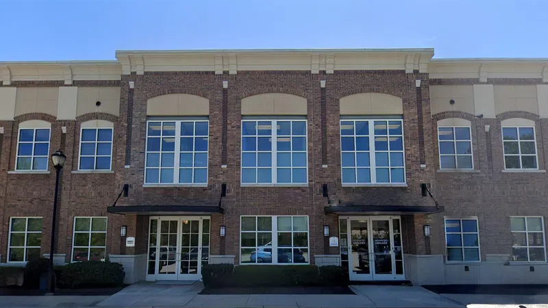 Brick office building with large windows, glass entrance doors, and trimmed shrubs under a clear blue sky.