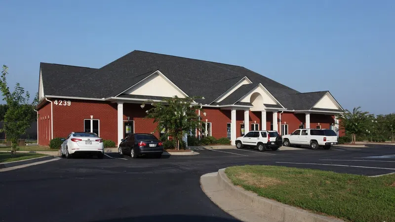 Red brick office building with white trim, black roof, parking lot, and several cars under clear blue sky.
