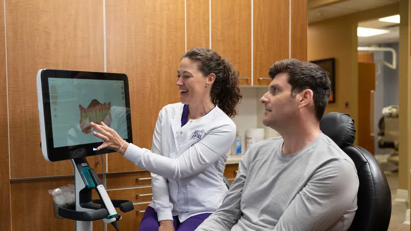 Dentist explaining 3D dental scan results to male patient in dental office with wooden cabinets.