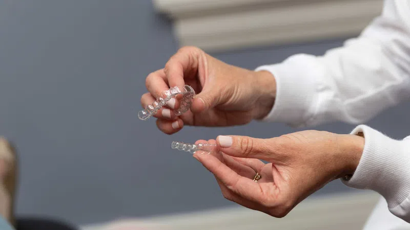 Close-up of hands holding clear dental aligners in a clinical setting with a patient in background.