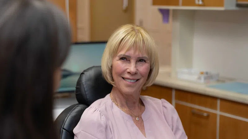 Smiling senior woman in pink blouse sitting in medical office talking to a healthcare professional.