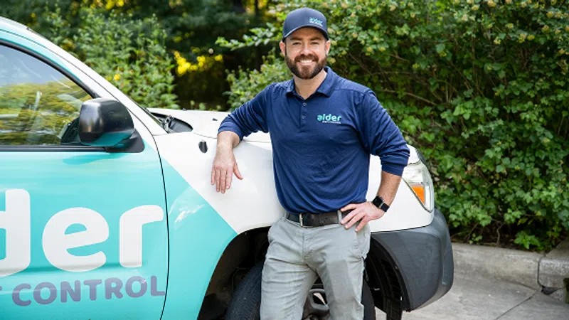 Smiling pest control technician in company uniform leaning on branded service vehicle outdoors