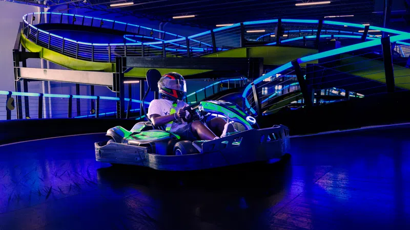 Person racing a green go-kart on an indoor track with blue and purple neon lights and safety railings.