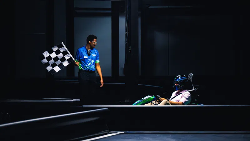Race official waves checkered flag to finish go-kart race with driver in helmet at indoor track.