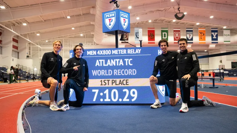 Four male athletes pose beside a digital scoreboard showing Atlanta TC's world record in men's 4x800 meter relay with time 7:10.29.