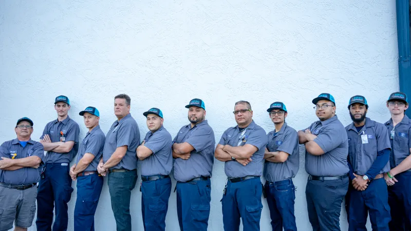 Group of eleven uniformed workers standing in a row against a white wall with arms crossed.