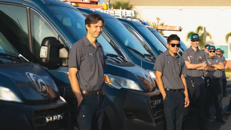 Technicians in uniform standing beside blue service vans lined up outdoors on a sunny day.
