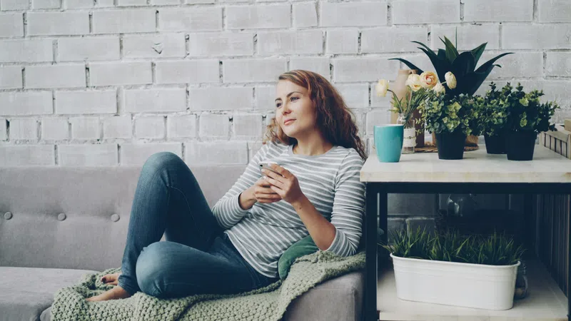 Young woman in striped shirt relaxing on a gray couch holding a smartphone with plants on a nearby table