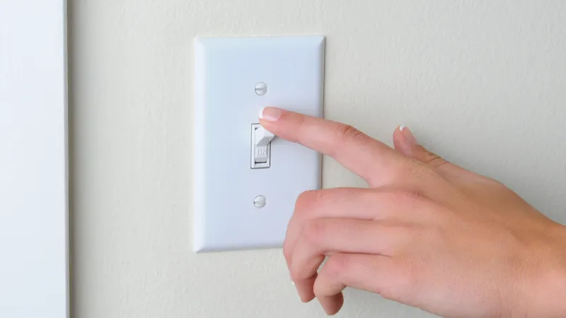 Hand with neatly manicured nails flipping a white wall light switch turned on against a beige wall.