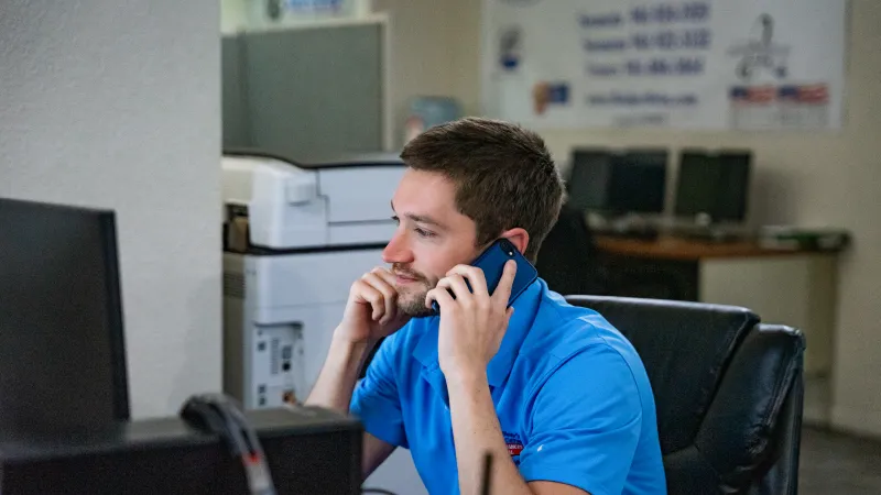 Man in blue shirt talking on phone at office desk with computer and printer in background