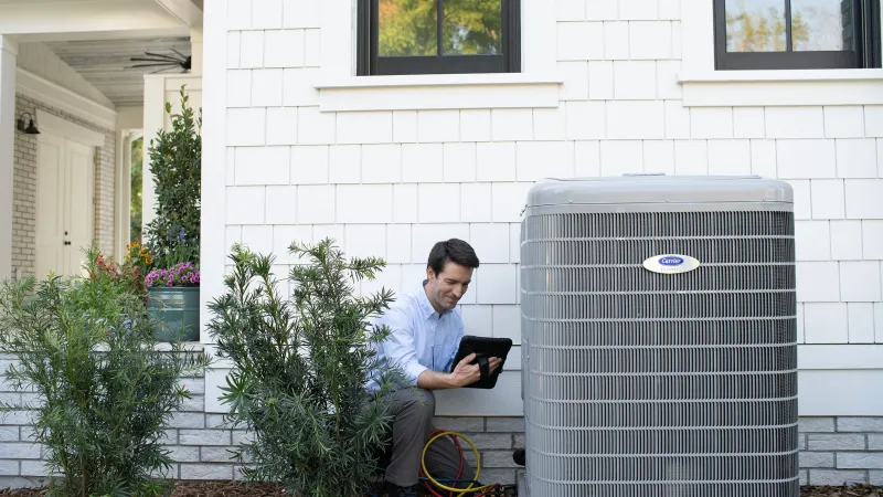 Man using a tablet while seated next to an air conditioning unit outside a home.