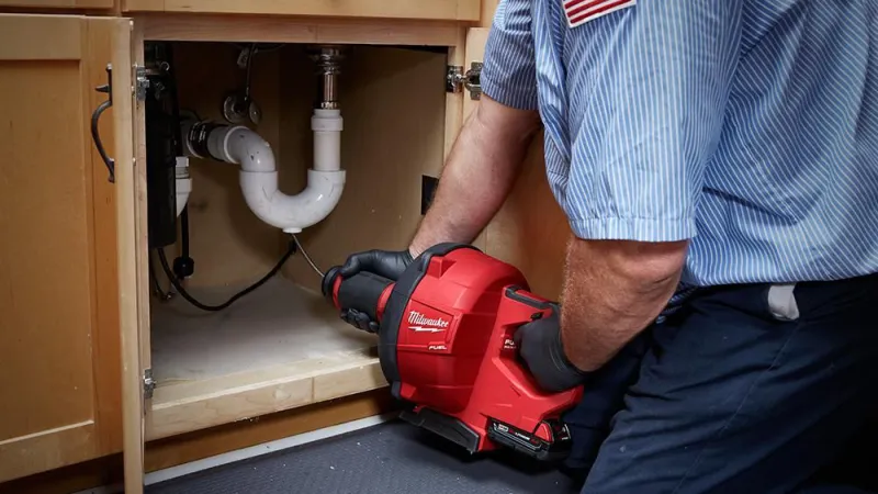 A technician using a Milwaukee drain auger to clear a sink blockage under a kitchen cabinet.