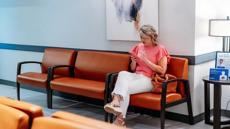 Woman wearing pink top and white pants sitting on brown leather bench in modern waiting room using smartphone