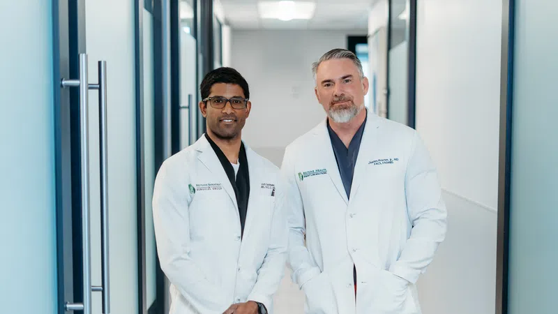 Two male doctors in white coats standing in a modern medical clinic hallway, smiling at the camera