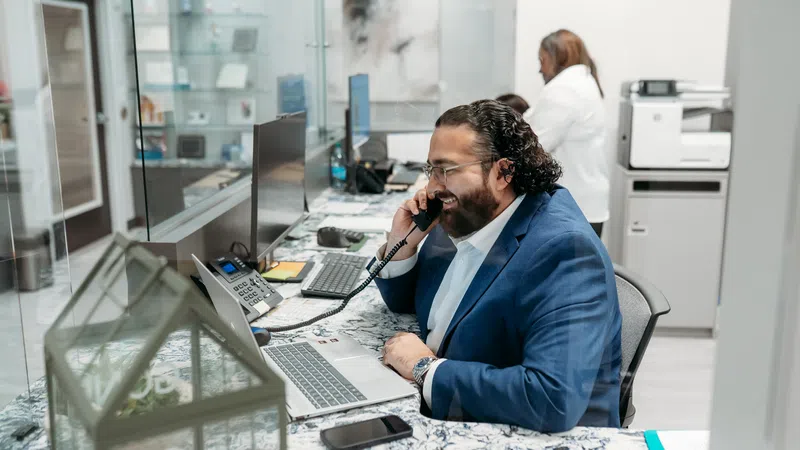 Man in blue suit talking on office phone while working on laptop in modern office environment.
