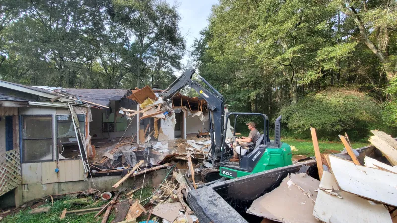 Man operating excavator to demolish a wooden house surrounded by trees with debris scattered around.