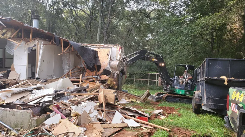 Mini excavator demolishes a small wooden structure amid a forested backyard with debris and trucks nearby