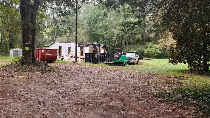 Small excavator and truck near white building under demolition in wooded rural yard with dirt ground and trees.