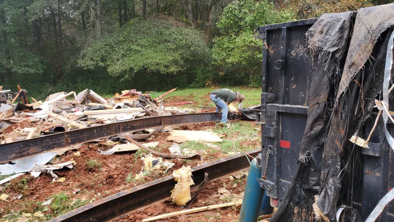 Worker welding metal beams on a muddy construction site surrounded by debris and trees in the background