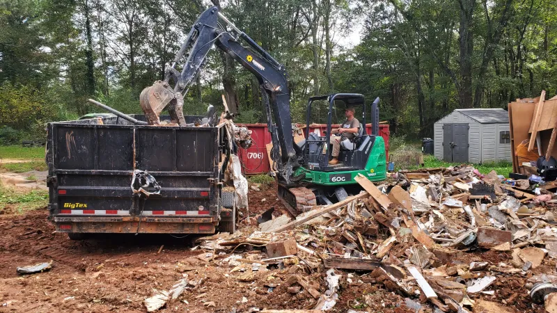 Excavator loading debris of demolished wood and construction materials into a dumpster at a clearing site