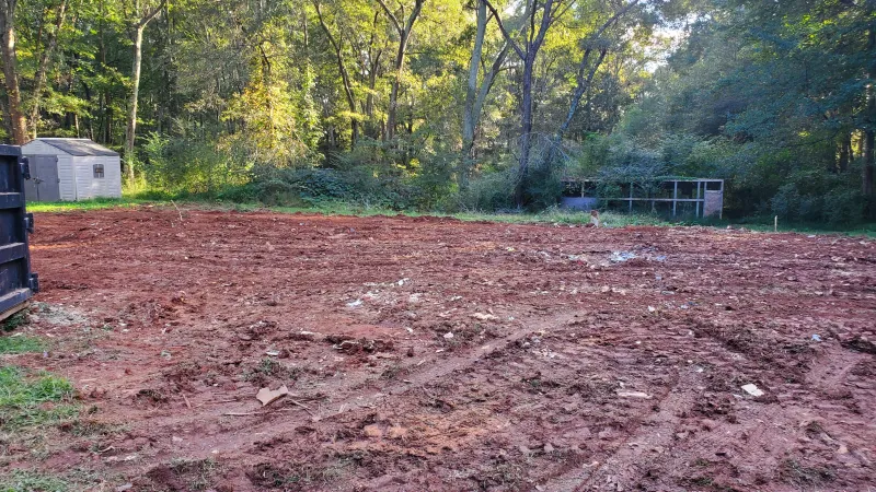 Cleared plot of red dirt land with tire tracks surrounded by trees and small outdoor sheds in the background.