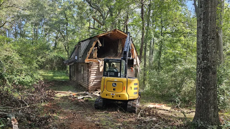 Yellow excavator demolishing a wooden cabin surrounded by dense green forest and trees on a sunny day