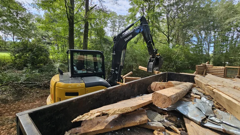 Excavator loading wooden logs and debris into a large dumpster on a sunny day in a forested area.