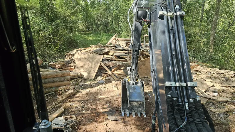Excavator clearing debris and wood piles in a forested area on a sunny day