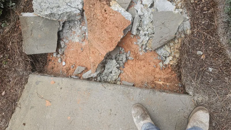 Broken concrete sidewalk with exposed red dirt and person standing on intact edge wearing work boots and jeans.