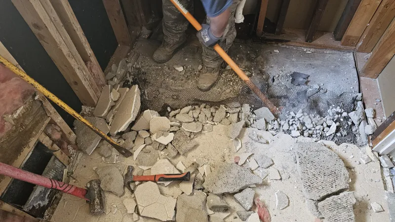 Worker breaking up concrete floor with tools during home renovation in an unfinished room.
