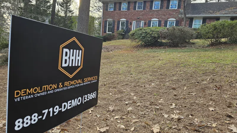 BHH demolition and removal services sign in front of a large brick house with shrubs and trees.