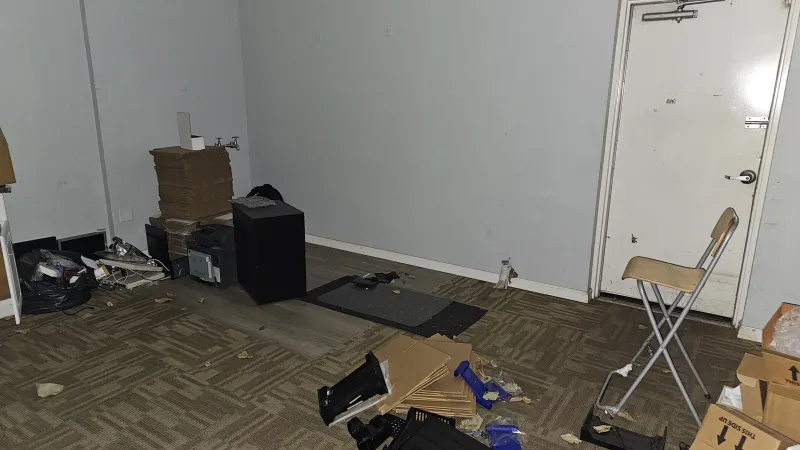 Cluttered storage room with scattered boxes, bags, equipment, and a folding chair near an exit door.