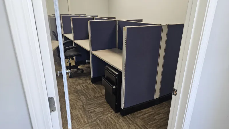 View into an office with empty cubicles and swivel chairs under neutral lighting and beige walls.
