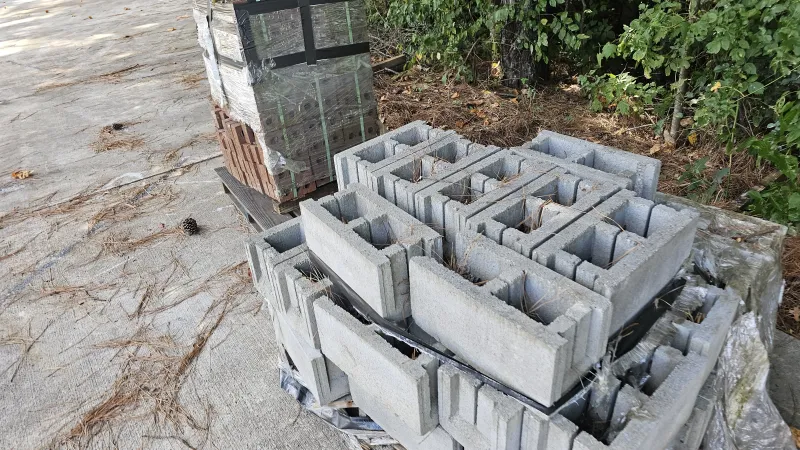 Stacked concrete blocks and bricks on pallets outdoors near wooded area and paved surface.