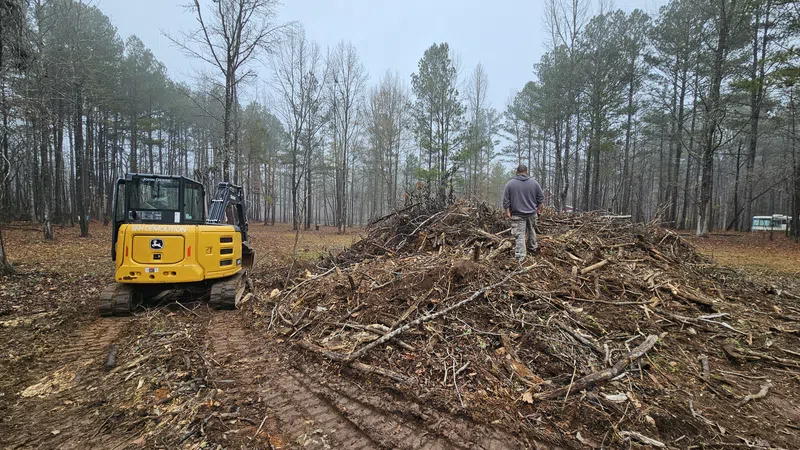 Man standing on large pile of branches and debris near yellow excavator in forest clearing on foggy day
