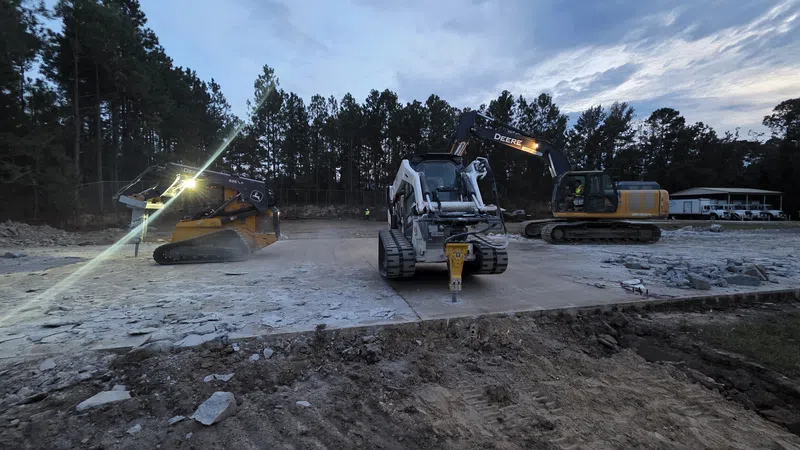 Three excavators working on a construction site at dusk with rubble and dirt in the foreground and trees in the background.