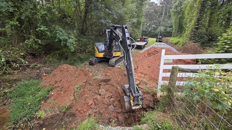 Small excavator digging a trench near a fence and wooded area for construction or drainage work.