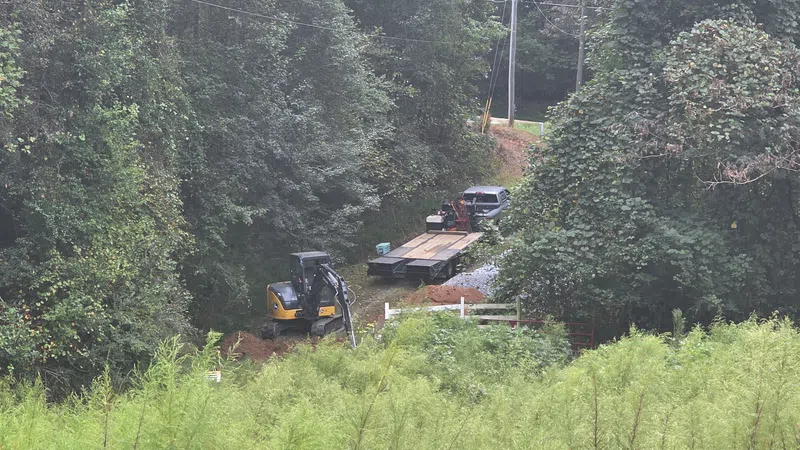 Construction site with excavator and flatbed truck in a forested area surrounded by dense green foliage.