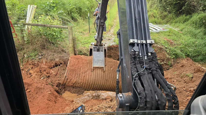 Excavator installing a large corrugated metal pipe in a muddy trench surrounded by green vegetation.