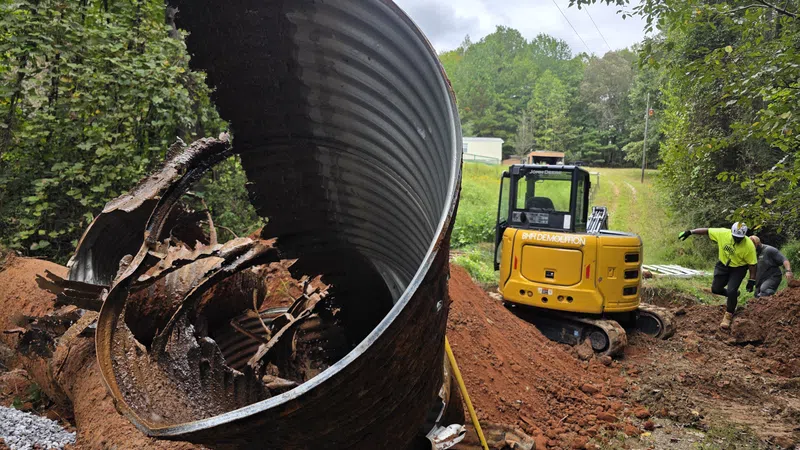 Large rusted metal culvert pipe removed from a trench with a yellow mini excavator and workers in a wooded area.