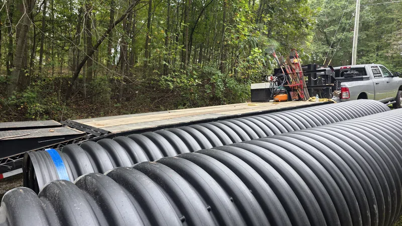 Large black corrugated drainage pipes stacked near a pickup truck and forested area ready for installation.