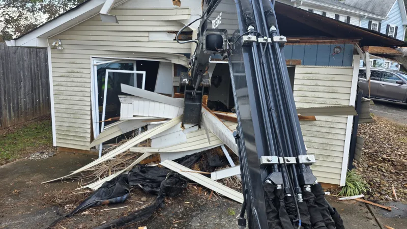 Excavator arm demolishing a small white building with broken siding and debris scattered around.