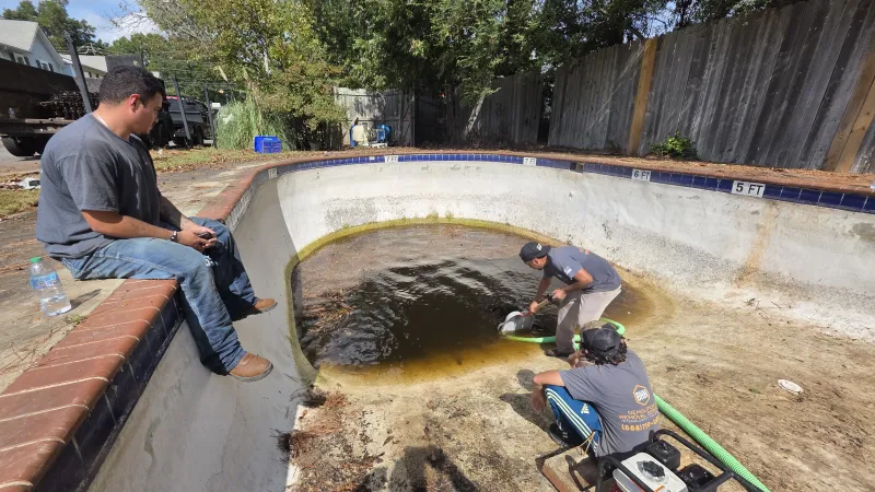 Three men working to pump out dirty water from an empty backyard swimming pool on a sunny day.