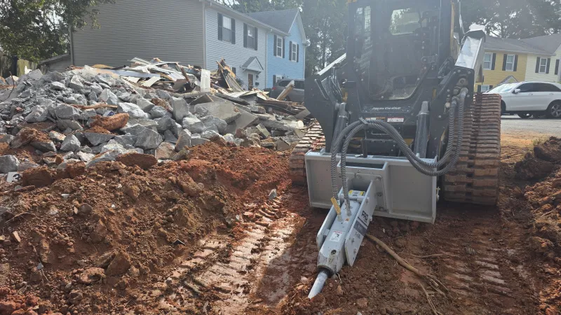 Tracked excavator with hydraulic breaker working on soil near demolished house rubble in residential area.