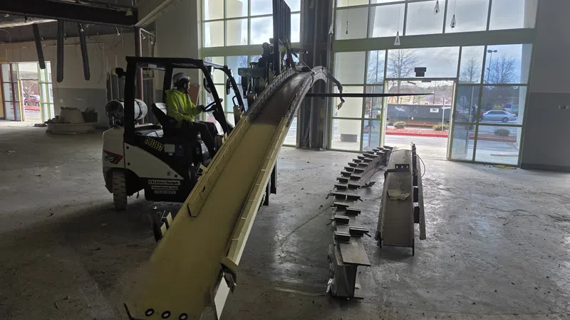 Worker operating forklift lifting a large curved metal beam inside a spacious industrial warehouse.
