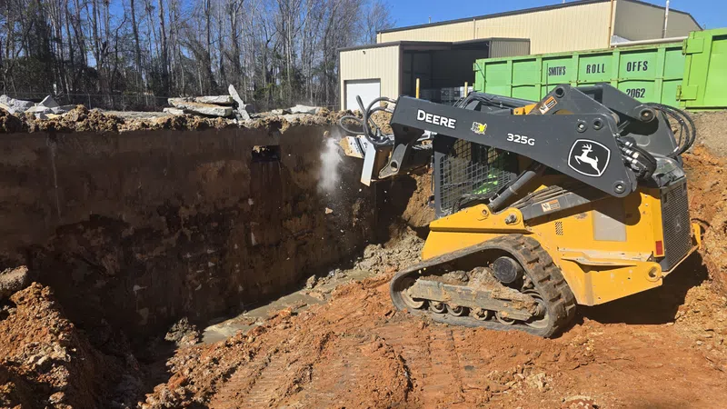 John Deere 325G compact track loader excavating soil at a construction site with a green dumpster in the background