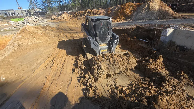 Compact bulldozer moving soil at a construction site surrounded by dirt mounds and forest trees.