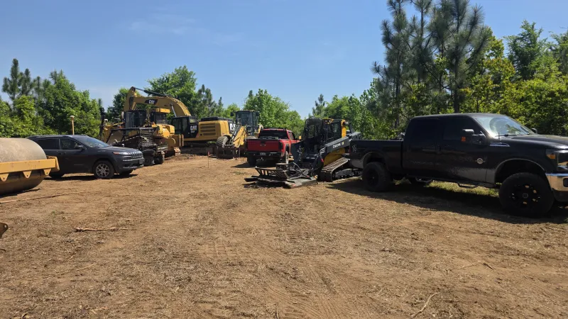 Construction vehicles and trucks parked on a dirt lot surrounded by green trees under a clear blue sky