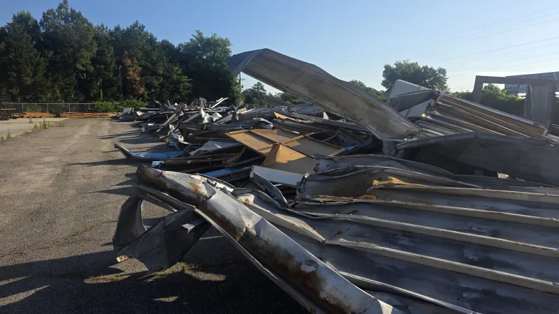 Piles of twisted and dented metal sheets lying in an outdoor lot with trees and clear blue sky.