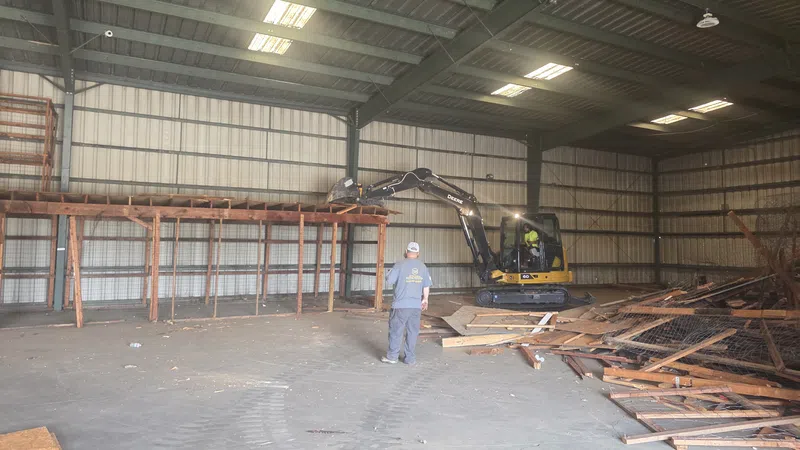 Worker operating excavator inside warehouse clearing wooden debris from metal shelving demolition.